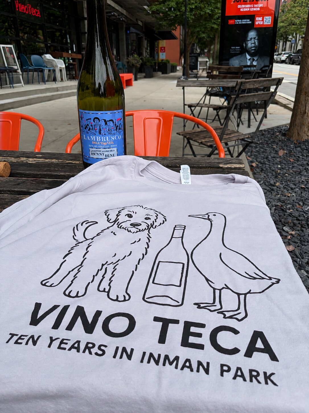 A lavender tenth-anniversary t-shirt featuring a fluffy dog, wine bottle, and duck, displayed in front of the VinoTeca storefront
